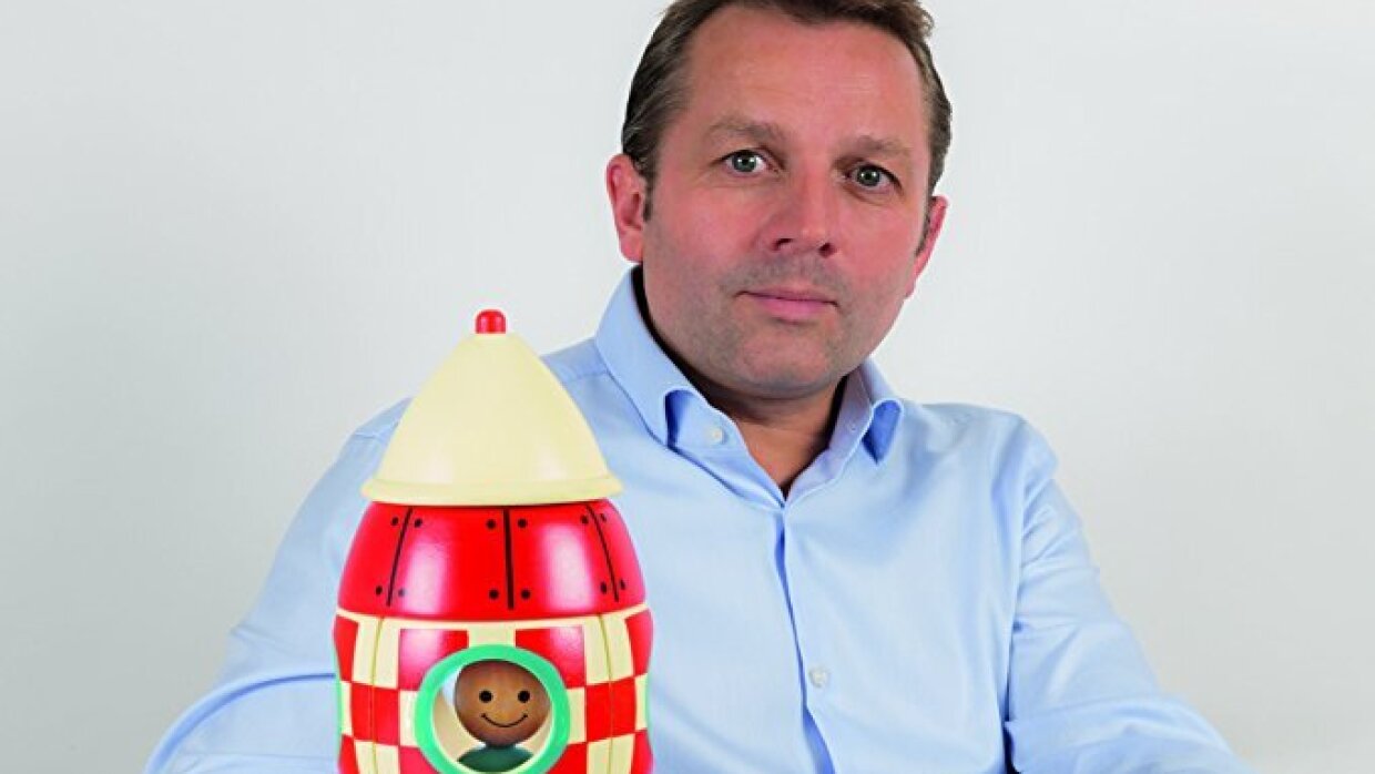 Le fondateur de Janod se tient devant l'un de ses jouets en bois qui représente une fusée à carreaux rouges et blancs avec un dessin naïf de personnage regardant par un hublot