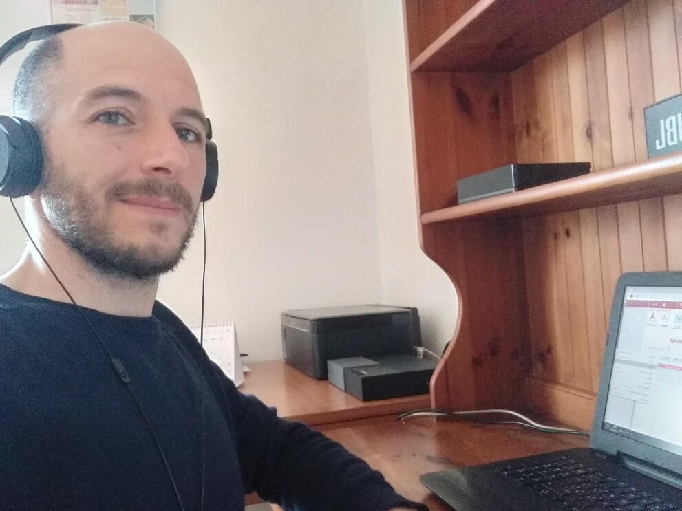 Primo piano di un uomo sulla sinistra. Indossa una maglia nera e delle cuffie, siede di fronte a una scrivania su cui è posato un PC. 