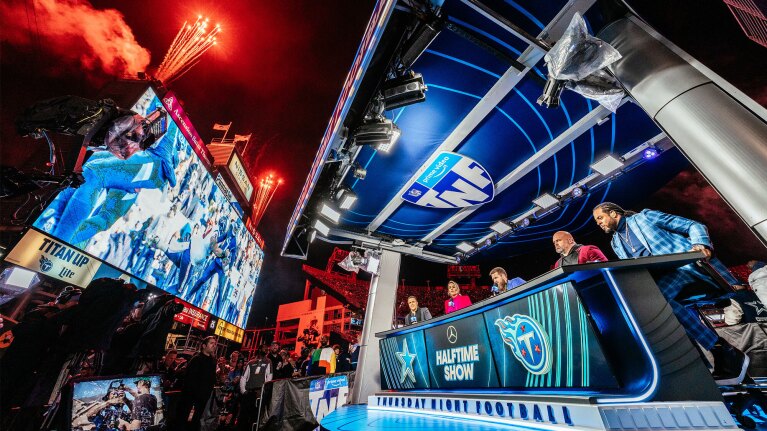 An image of the Thursday Night Football hosts sitting at the announcing table on the field. There is a large, blue screen in front of them and red fireworks taking flight in the air above.