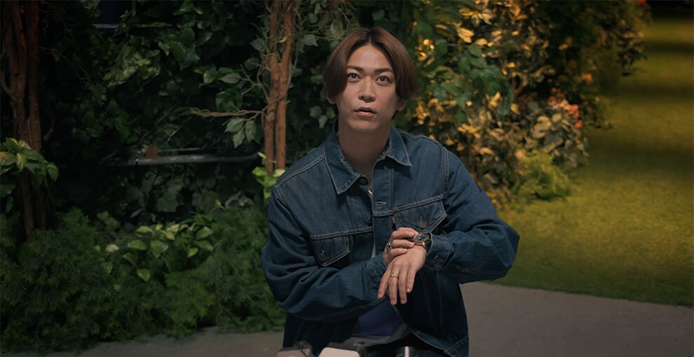 緑の植栽と人工芝の薄暗い背景に、インディゴブルーのデニムジャケットを着た人物が立っている。上方を見上げるポーズで、胸の高さで左手の腕時計に右手を添えている。