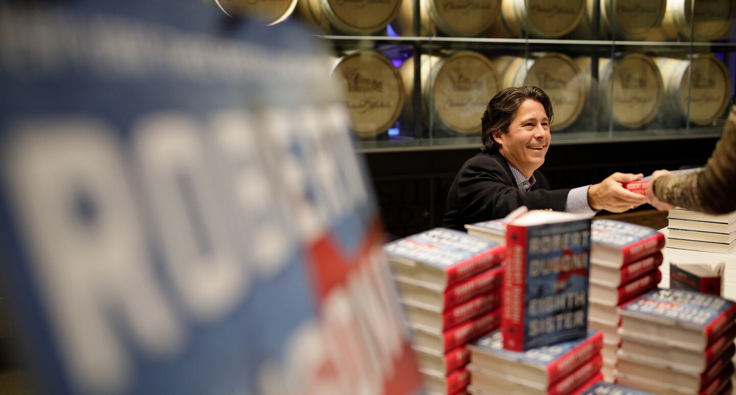 A man sits at a table covered with stacked copies of books. He hands a book another person.