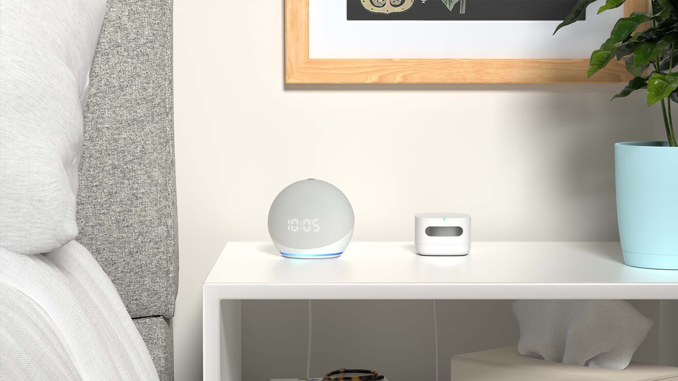 An Alexa Echo device sits on a nightstand next to a bed.