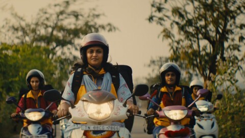 Cuatro mujeres indias en moto. Todas ellas llevan casco y una mochila grande donde cargan los pedidos. Las motos son dos de color blanco y dos de color morado y van con las luces endendidas. De fondo árboles.  y un cielo de color marrón.