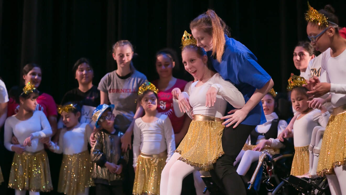 A dancer wearing ballet slippers, a gold skirt, a tiara, and a white leotard and tights performs. The dancer has cerebral palsy and is leaning back against a helper for support. Other dancers and helpers are in the background.
