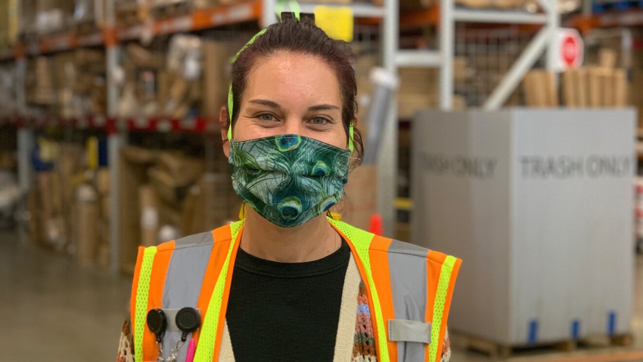 A woman wearing a safety vest and a mask that covers her mouth and nose stands in a space with tall shelves in the background.