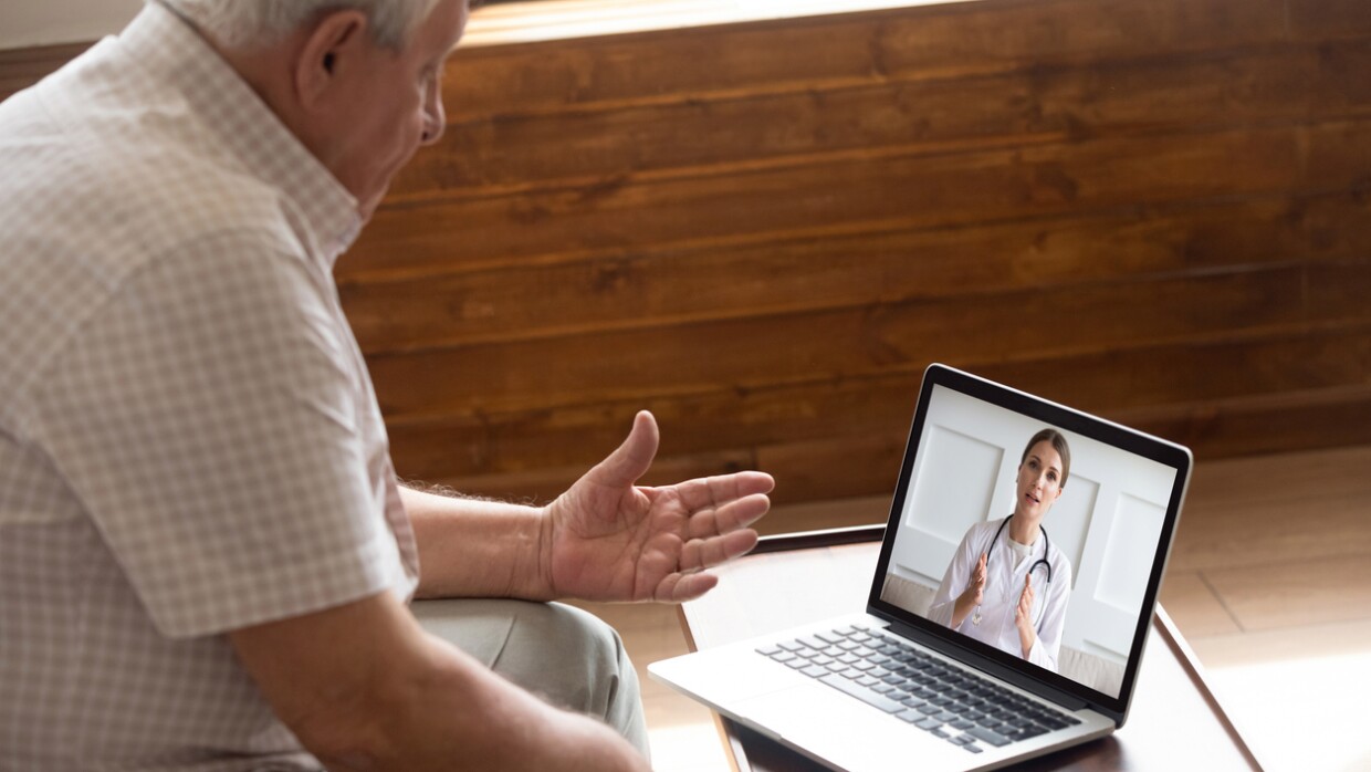 A man sits in front of his computer, which rests on a side table. On the screen of the laptop computer is a woman wearing a lab coat and stethescope, appearing to speak to him.