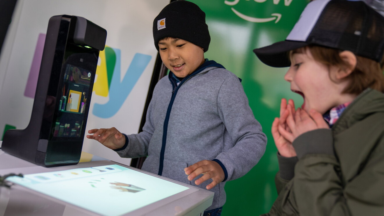 An image of two boys smiling while playing with the display version of Amazon Glow at an event. The device is projecting an interactive screen on a table and they are playing  the games and activities on it.