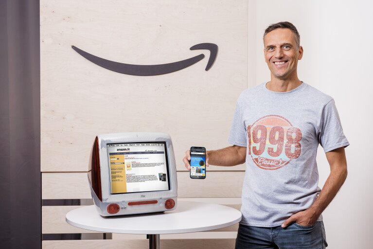 Amazon Deutschland gibt es seit 25 Jahren. Rocco Bräuniger hält ein Smartphone mit der Amazon App vor einen PC mit der Amazon Website.