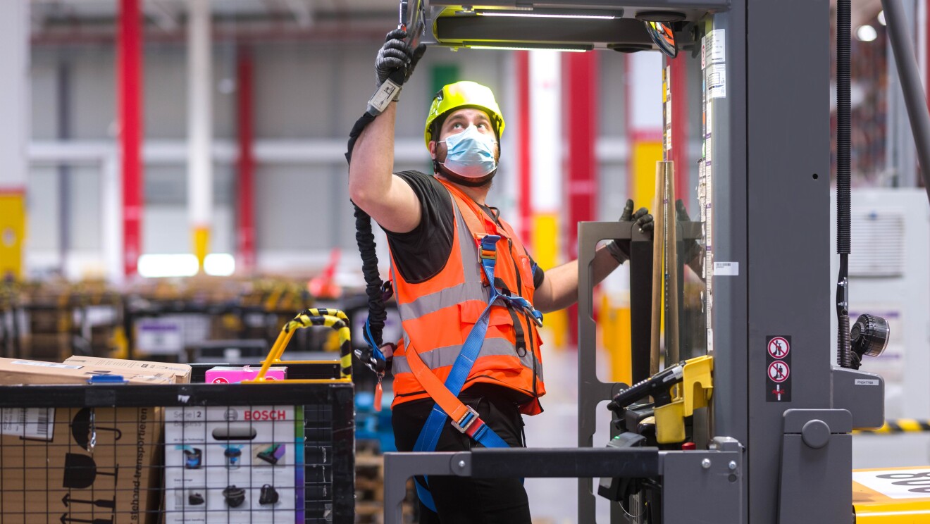 Dipendente di un magazzino Amazon. Indossa un casco, una mascherina chirurgica e un giubbino di sicurezza, si trova al centro di un macchinario. Alle sue spalle un cesto con dei pacchi Amazon.