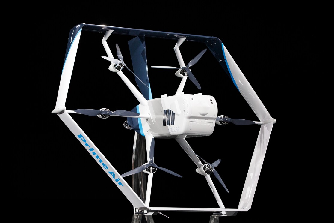 A white and light blue Prime Air drone in front of a black background. Six propellers and a main body are visible in the center of the drone.