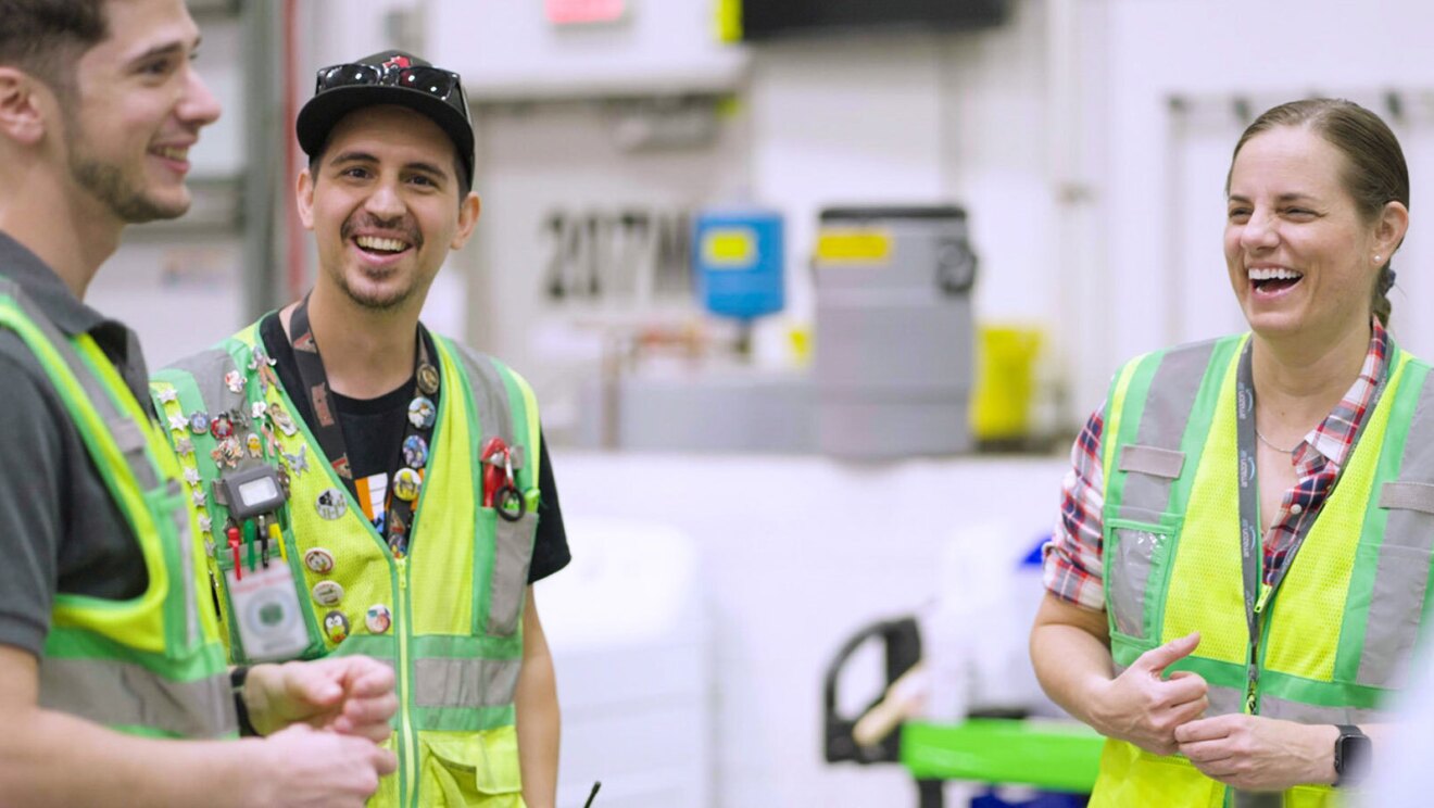 An image of Sarah Rhoads standing inside a fulfillment center with two other Amazon employees talking and laughing.