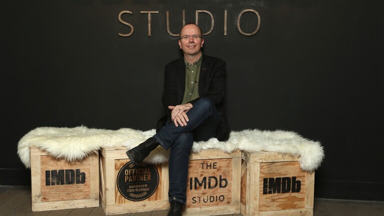 Cal sitting on IMDb branded boxes with a white rug on top. There is a sign behind him that reads "studio."