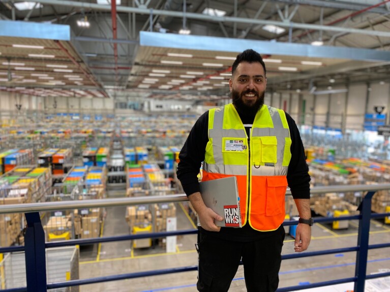 Spezialist für Arbeitssicherheit bei Amazon Mustafa Almujahed steht mit einer gelb-orangenen Weste bekleidet samt Laptop in einem Logistikzentrum.
