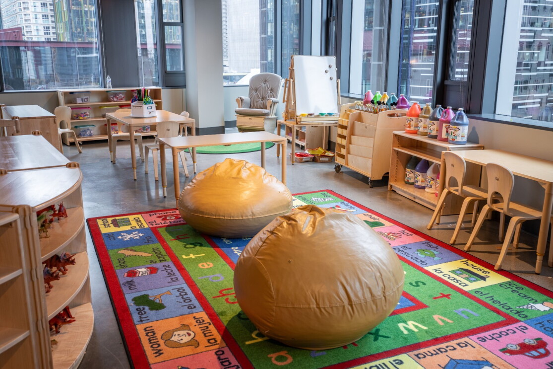 An image of a room with space for kids to play. There is a colorful mat on the floor, bean bags, other chairs, and toys for kids to play with.