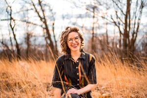 A professional image of Katie (schlosser) vitolins, head of Amazon Music for Artists. She is smiling for a phot sitting in a field. 