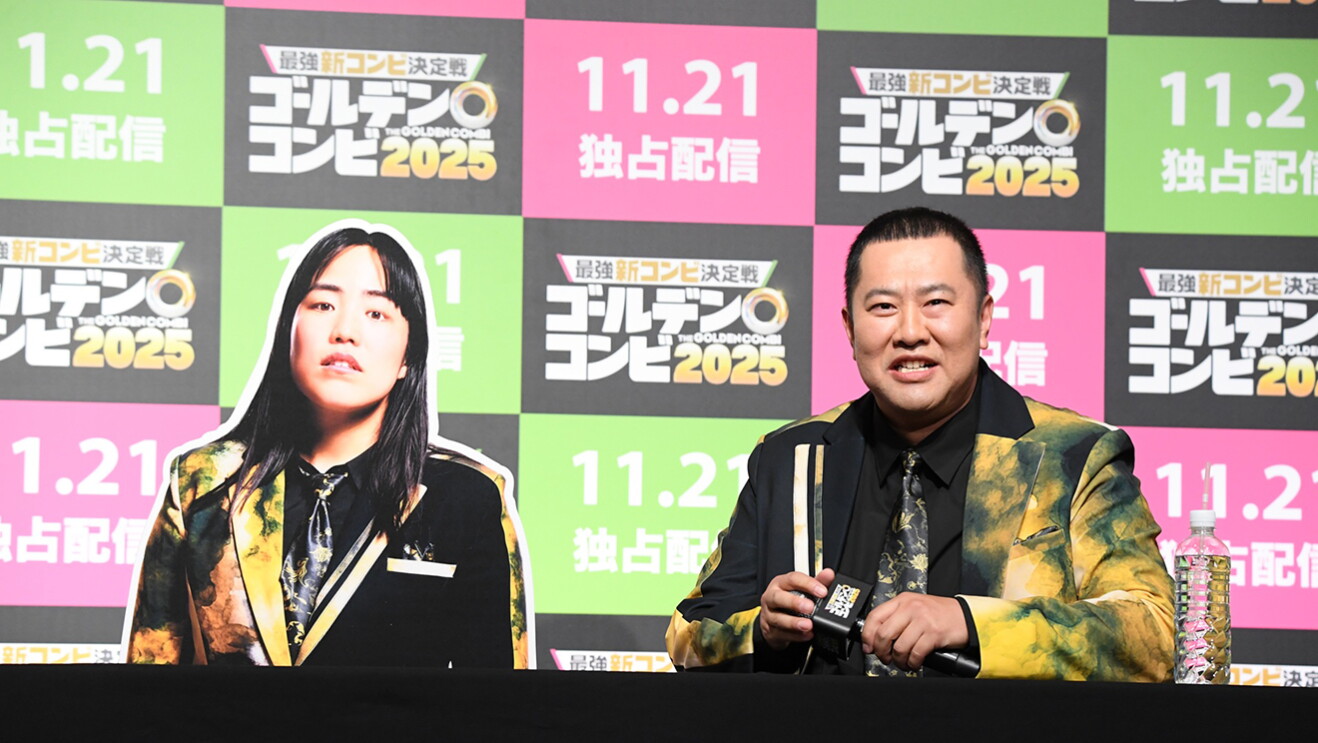 ピンク、黄緑、黒の市松模様の壁を背景に、マイクを持って着席した男性。黒と黄色マーブルの派手なスーツ衣装を着用。男性の左側に、同じ衣装姿の女性のパネルが置かれている。