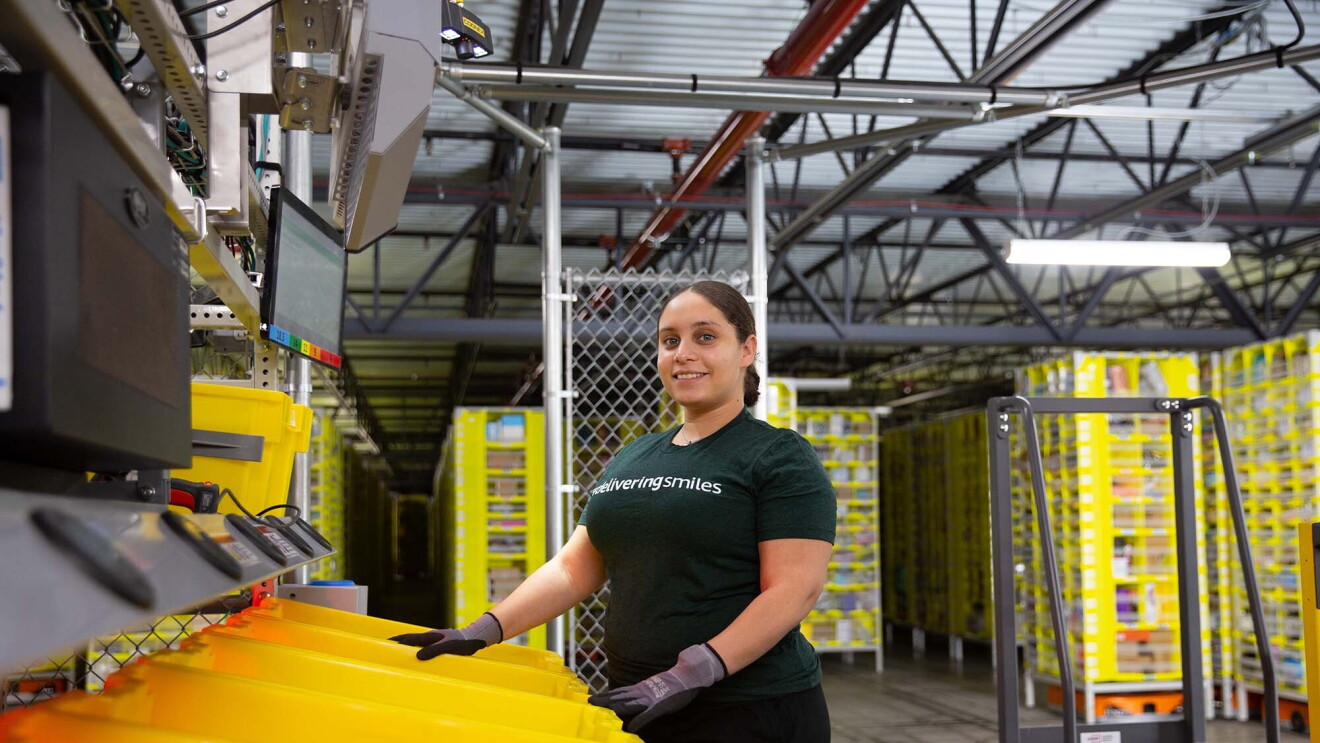 A woman working at an Amazon fulfillment center stands in front of a row of yellow bins. 