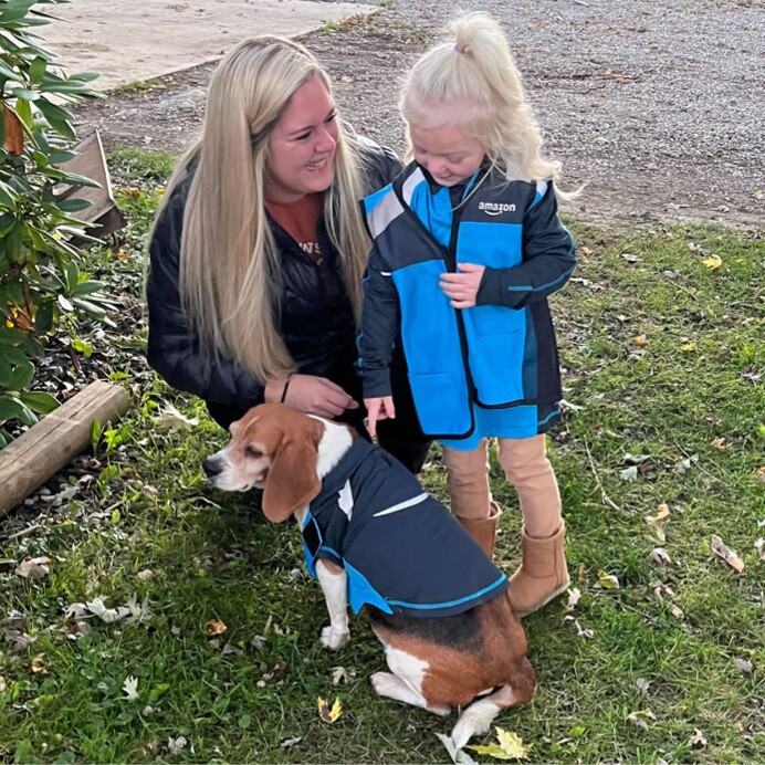 Oaklee wears an Amazon delivery driver costume and stands next to her mom who is crouching down and smiling at Oaklee. Oaklee is pointing at her dog who is also dressed up in an Amazon delivery driver costume.