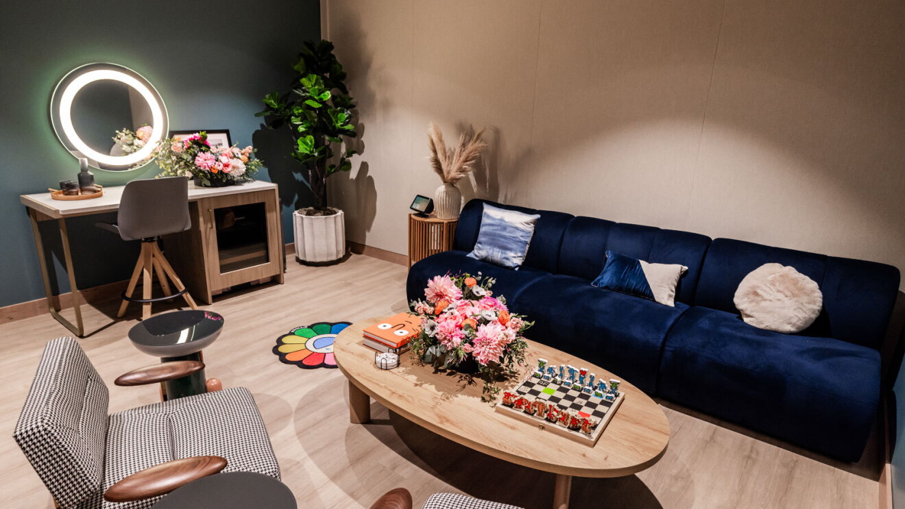 A room with tan and teal accents. There is a dark blue couch, tan chairs, a small, colorful flower rug, and a vanity with a backlit mirror.