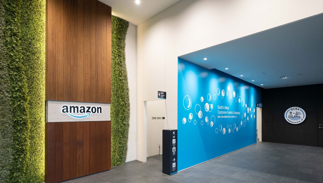 左側は、天井まで続く壁面植物で飾られた木目の壁面。木目の壁面にAmazonのロゴ。右側に白い壁と奥に続く通路。通路の壁は水色で、シャボン玉のようなイラストと英文が白でかかれている。