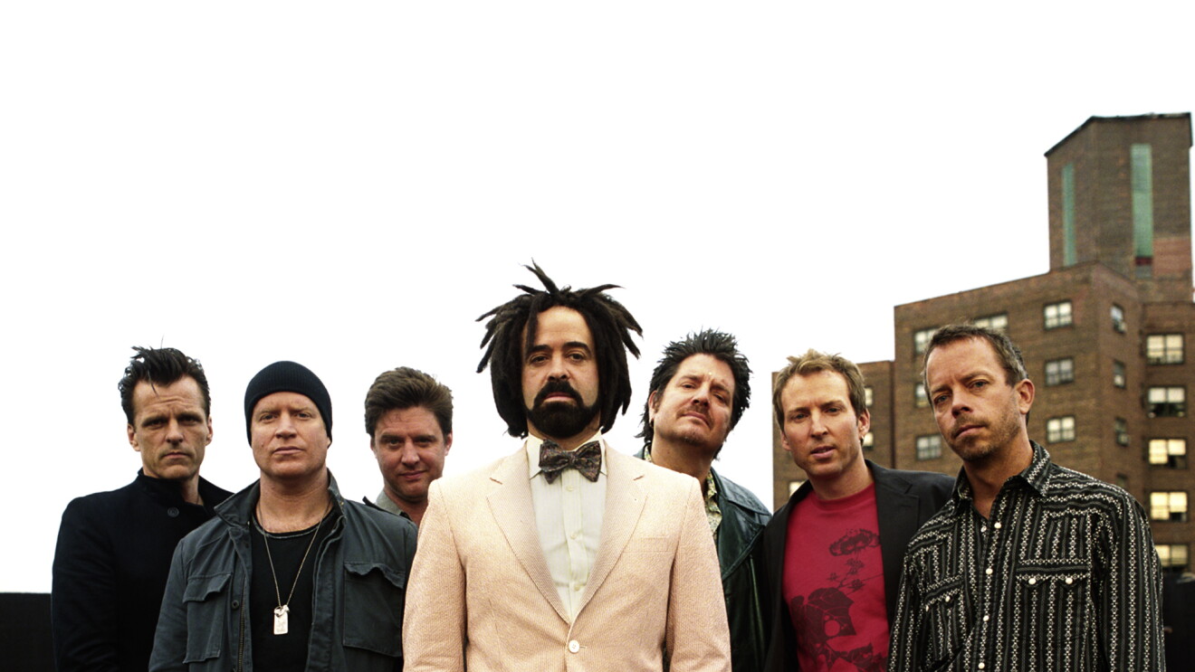 The Counting Crows band, six men stand together wearing casual clothing. In front of them, Adam Duritz stands in a cream suit with bow tie and tennis shoes.