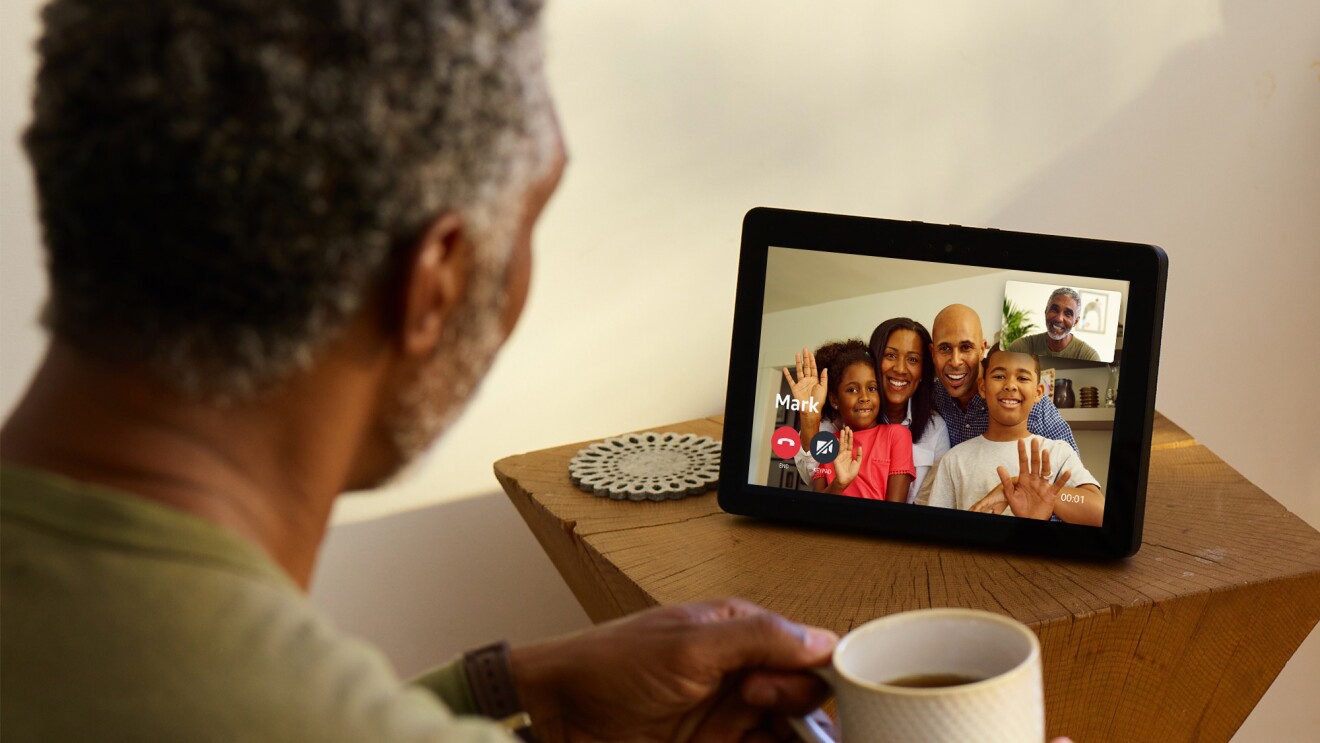 木制边桌上的Echo Show设备。在屏幕上,一家四口正微笑着挥手。面对设备的是一个拿着咖啡杯的男人。屏幕上显示了他的笑脸。