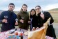 El equipo de Iberic Box, 4 jóvenes que no superan los 40 años, están posando detrás de un mesa lleva de comida y brindan con copas de vino tinto. En la izquierda, el CEO de la startup, David González, con una copa de vino tinto y un sobre de jamón, acompañado de tres colegas del equipo. Todos visten con chaquetas de invierno. La mesa tiene un mantel de cuadros, un jamón con el jamonero, un plato de jamón, las cajas de Iberic Box, una botella de aceite y otra de vino. El fondo de la foto es el cielo azul. 