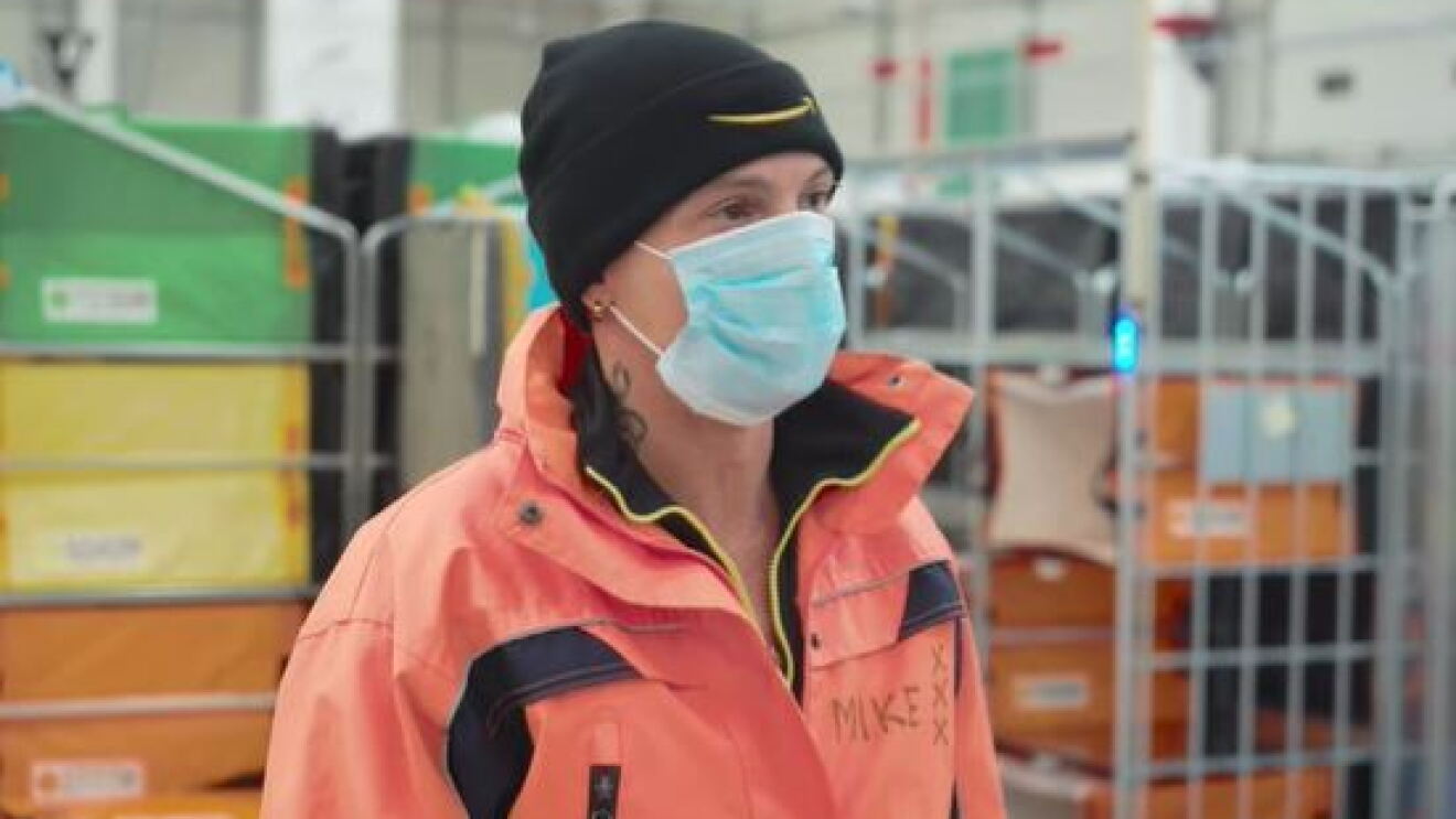Primo piano di un dipendente Amazon. Indossa un cappellino nero con lo Smile di Amazon ricamato, una mascherina chirurgica sul volto e un giubbotto arancione, con la scritta 'Mike' all'altezza del petto, a destra. 