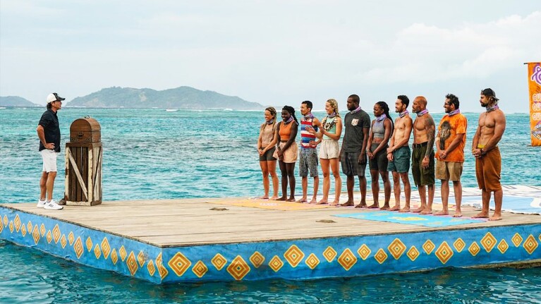 An image from the show Survivor of people standing on a pier with a person standing in front of them. There is water around them and small mountains in the background.