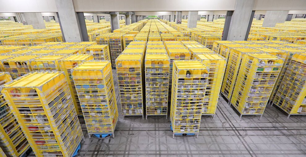 グレーの広大な物流センター内。整然と並ぶ黄色い収納棚の列が、視界のいちばん奥まで続いている。