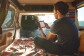 A man reading from a Kindle Paperwhite sits in the back of a camper van, in front of a large body of water.