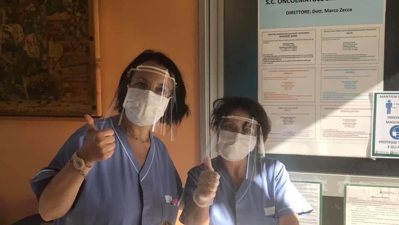 Due donne indossano una mascherina di plastica sugli occhi e una mascherina chirurgica sul volto. Guardano in camera e fanno segno 'OK' con il pollice verso l'alto. Di fronte a loro, alcune scatole di viveri.