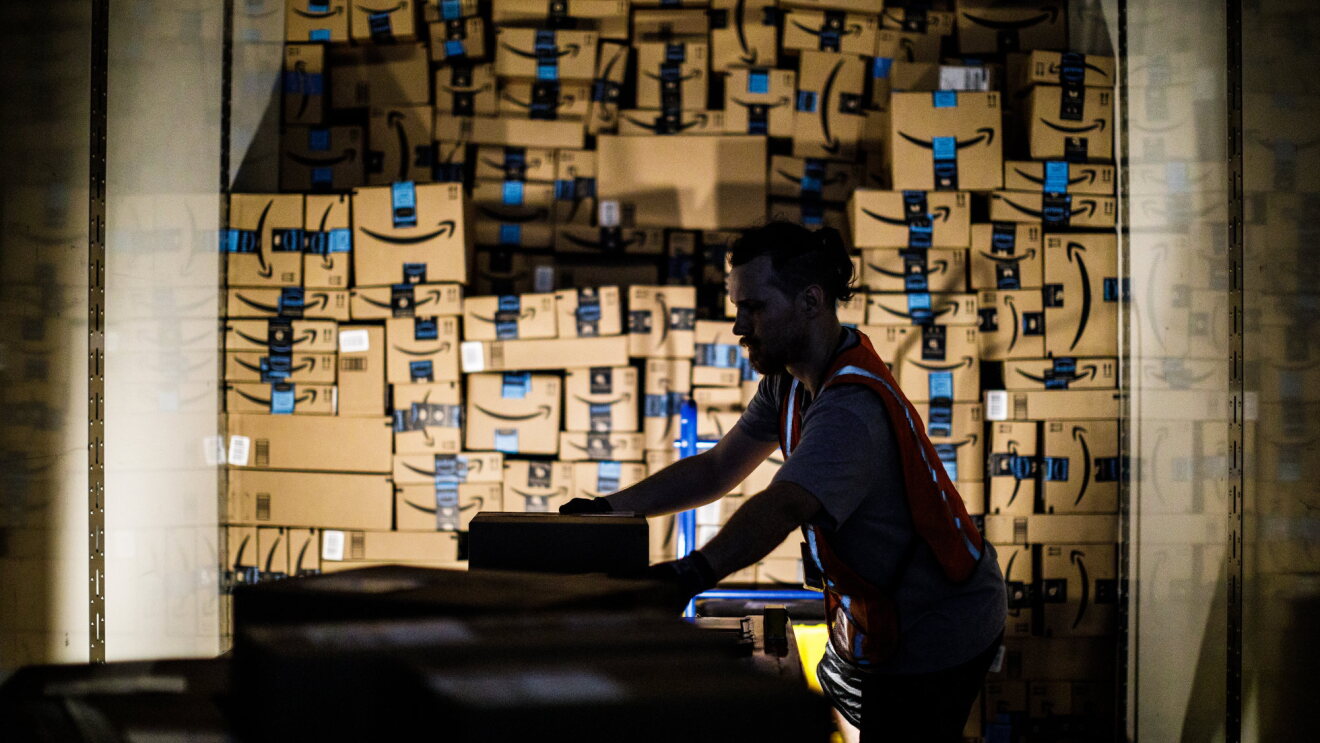 An Amazon associate, dramatically back lit by his work light, stacks shipping boxes labeled with the Amazon smile to the ceiling of a semi trailer.