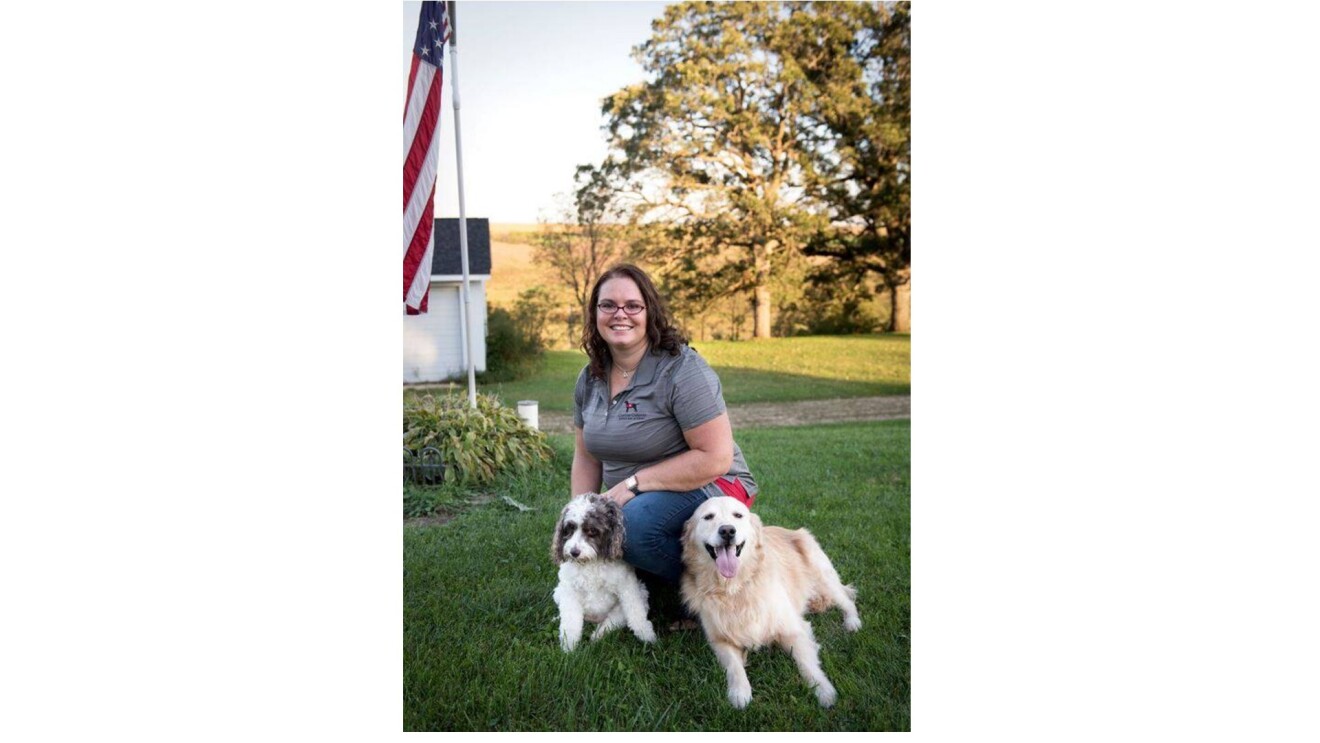 Shannon smiling and kneeling with two dogs by her side in a yard.