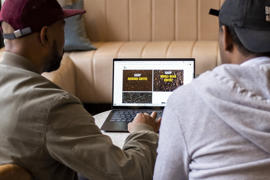 Two men sit side by side. The man on the left works on a laptop.