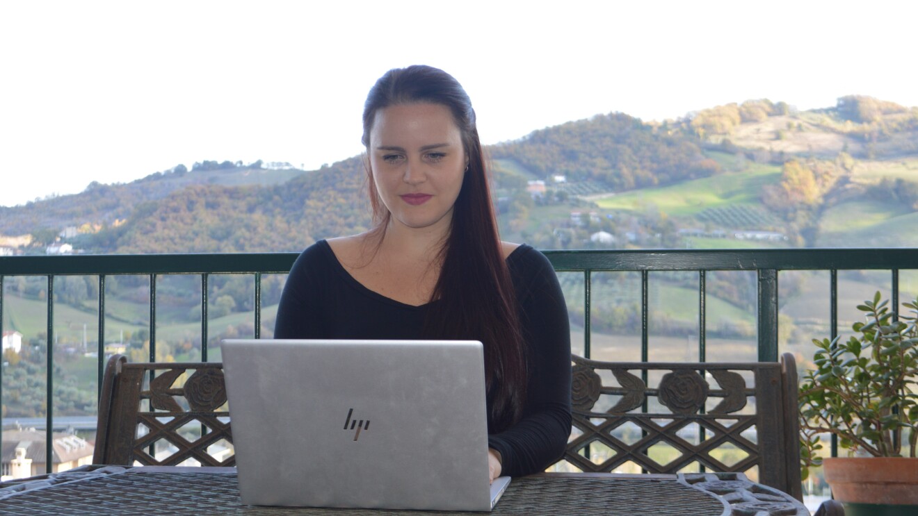 Una donna è seduta ad un tavolo su un terrazzo e ha di fronte a sè un PC. Alle sue spalle delle colline.