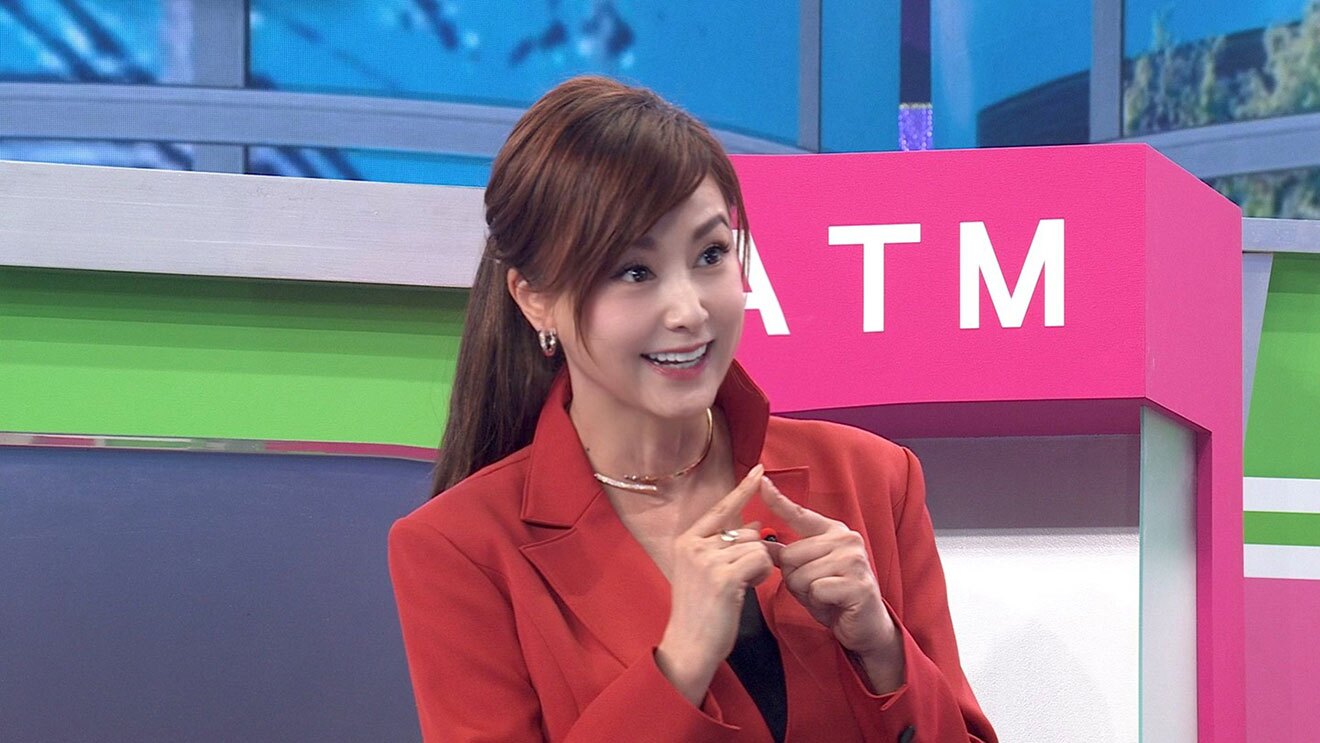 一部が黄緑やピンクに塗られたセットを背景に、赤い上着の女性の上半身。体の前で両手の人差し指を合わせ、笑顔を見せている。背後のピンクに塗られた部分に白字で「ATM」。