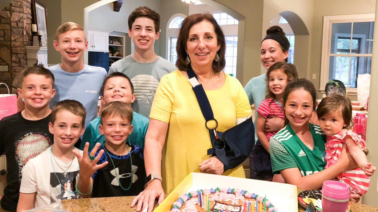 Rebecca en un cabestrillo que sostiene su brazo izquierdo está rodeada por sus diez nietos con un pastel de cumpleaños frente a ellos.