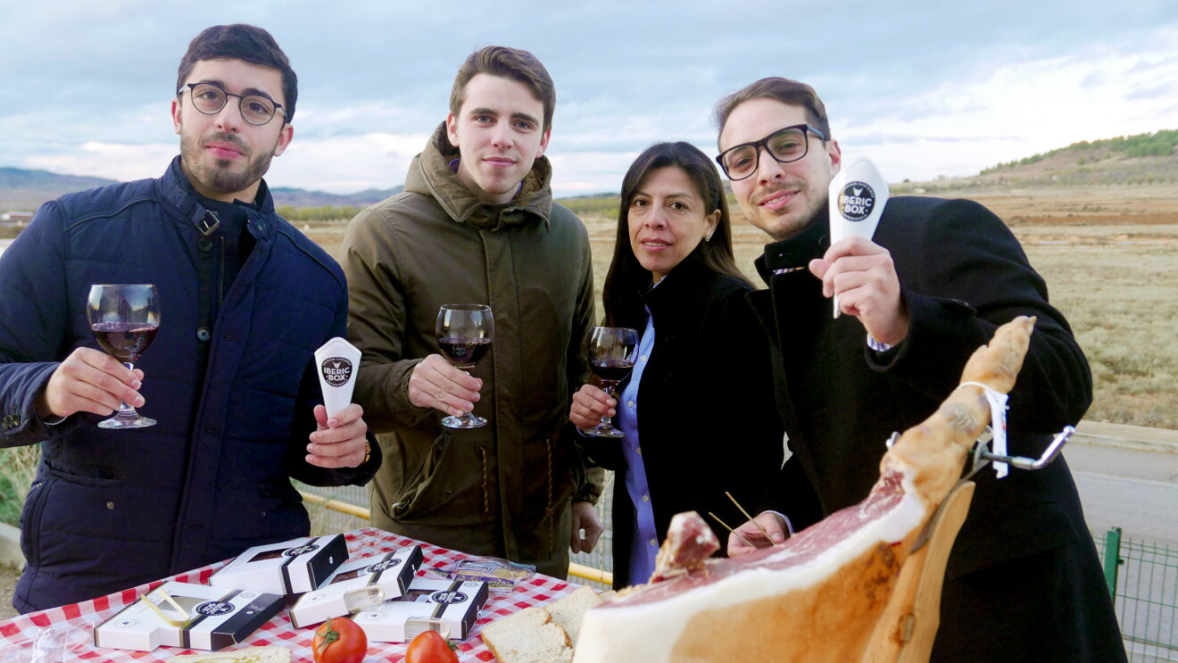 El equipo de Iberic Box, 4 jóvenes que no superan los 40 años, están posando detrás de un mesa lleva de comida y brindan con copas de vino tinto. En la izquierda, el CEO de la startup, David González, con una copa de vino tinto y un sobre de jamón, acompañado de tres colegas del equipo. Todos visten con chaquetas de invierno. La mesa tiene un mantel de cuadros, un jamón con el jamonero, un plato de jamón, las cajas de Iberic Box, una botella de aceite y otra de vino. El fondo de la foto es el cielo azul. 