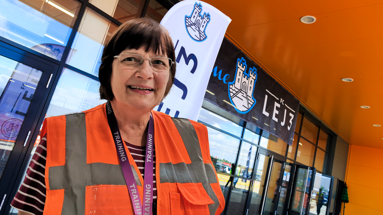 Eine Frau mit Brille und braunen Haaren, sie trägt eine orangene Sicherheitsweste. Im Hintergrund sieht man das Logistikzentrum.