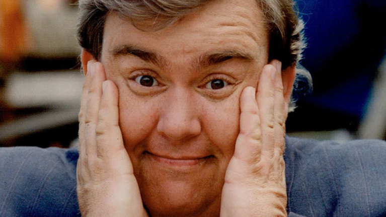 Actor John Candy smiling at the camera with hands on either side of his face
