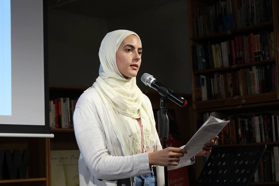 A woman (Sumaya Awad) stands in front of a microphone, speaking about her experience