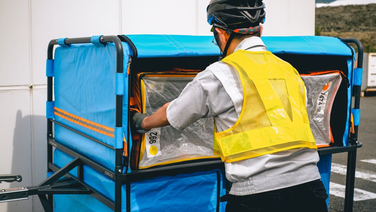 黄色い安全ベストを着てヘルメットをかぶっている男性が大きな荷物をリヤカーの中に積み込んでいる。