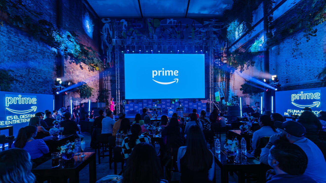 En una sala con una pantalla de fondo conel logo Prime aparecen mesas con personas sentadas mirando el escenario. Todo es de tonos de color azul. 