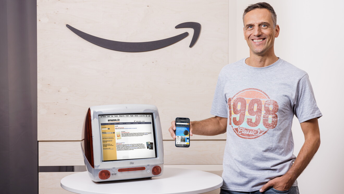 Amazon Deutschland gibt es seit 25 Jahren. Rocco Bräuniger hält ein Smartphone mit der Amazon App vor einen PC mit der Amazon Website.