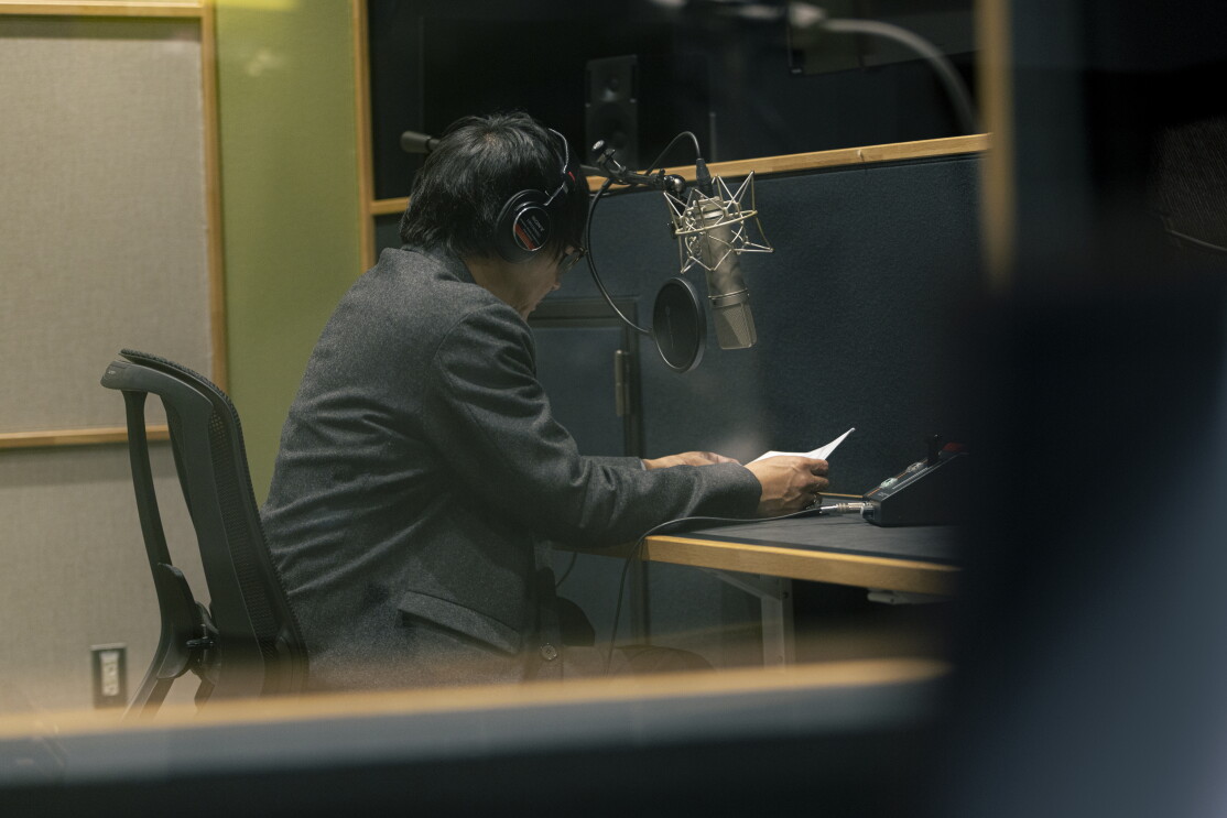 音声収録ブースをガラス窓ごしに見たところ。グレーのジャケットの男性がヘッドフォンをしてマイクに向かっている。