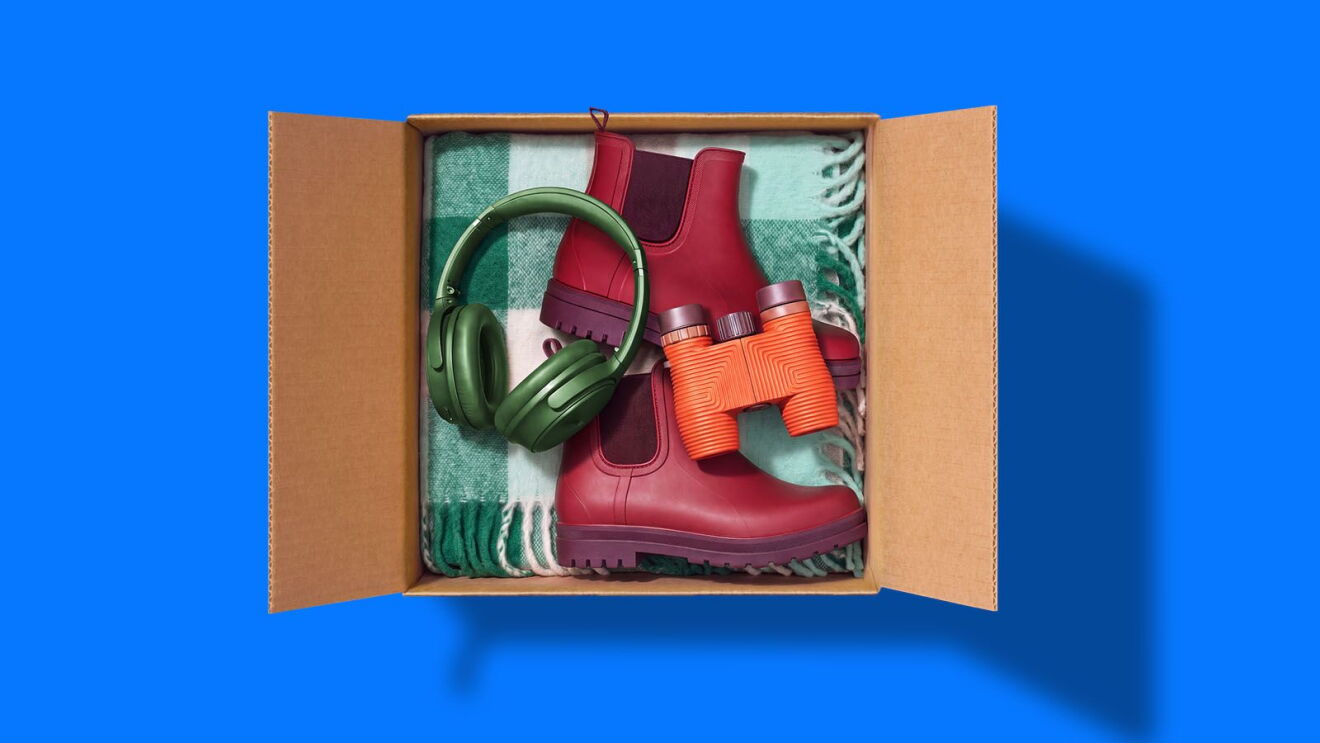 Open cardboard box with red rain boots, green headphones, orange binoculars, and a checkered blanket
