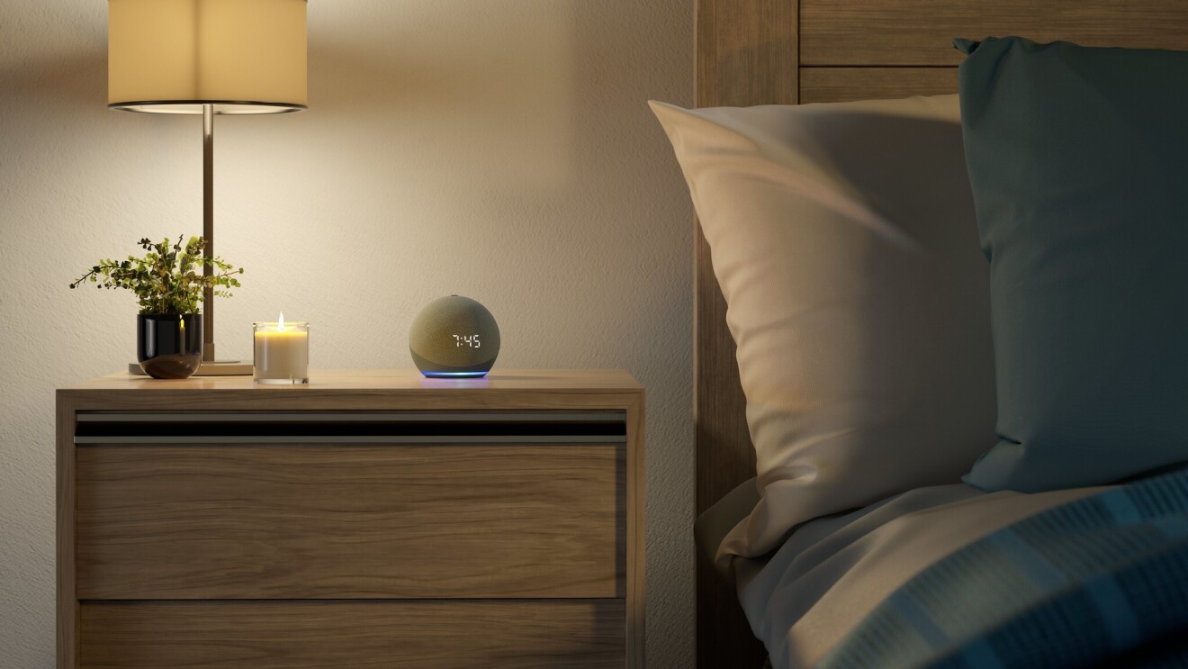 An Amazon Echo Dot on a nightstand next to a candle, a plant and a lamp.