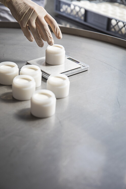 Auf einer grauen Arbeitsfläche stehen fünf geöffnete weiße Döschen, in die die handgemachte Deocreme Sommerliebe abgefüllt wurde. Am oberen linken Bildrand ist eine Hand mit Gummihandschuh zu sehen, die gerade ein sechstes Döschen auf eine Feinwaage gestellt hat. 