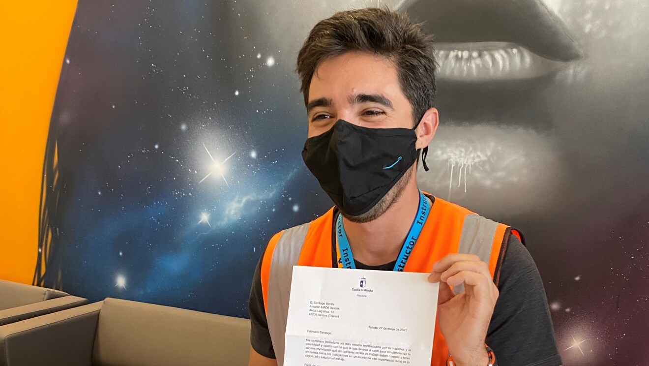 Santi Morilla sujetando la carta recibida del presidente de Castilla- La Mancha, emiliano García-Page. Con el pelo negro corto y una mascarilla negra con la sonrisa de Amazon en azul, lleva chaleco naranja y barba de dos días. Lleva camiseta negra de manga corta y unos jeans. Está apoyado en un sofá marrón de la recepción del centro logístico de Illescas (Toledo) y de fondo, en la pared, hay un poster con una sonrisa. Él aparece mirando a cámara y sonriendo.
